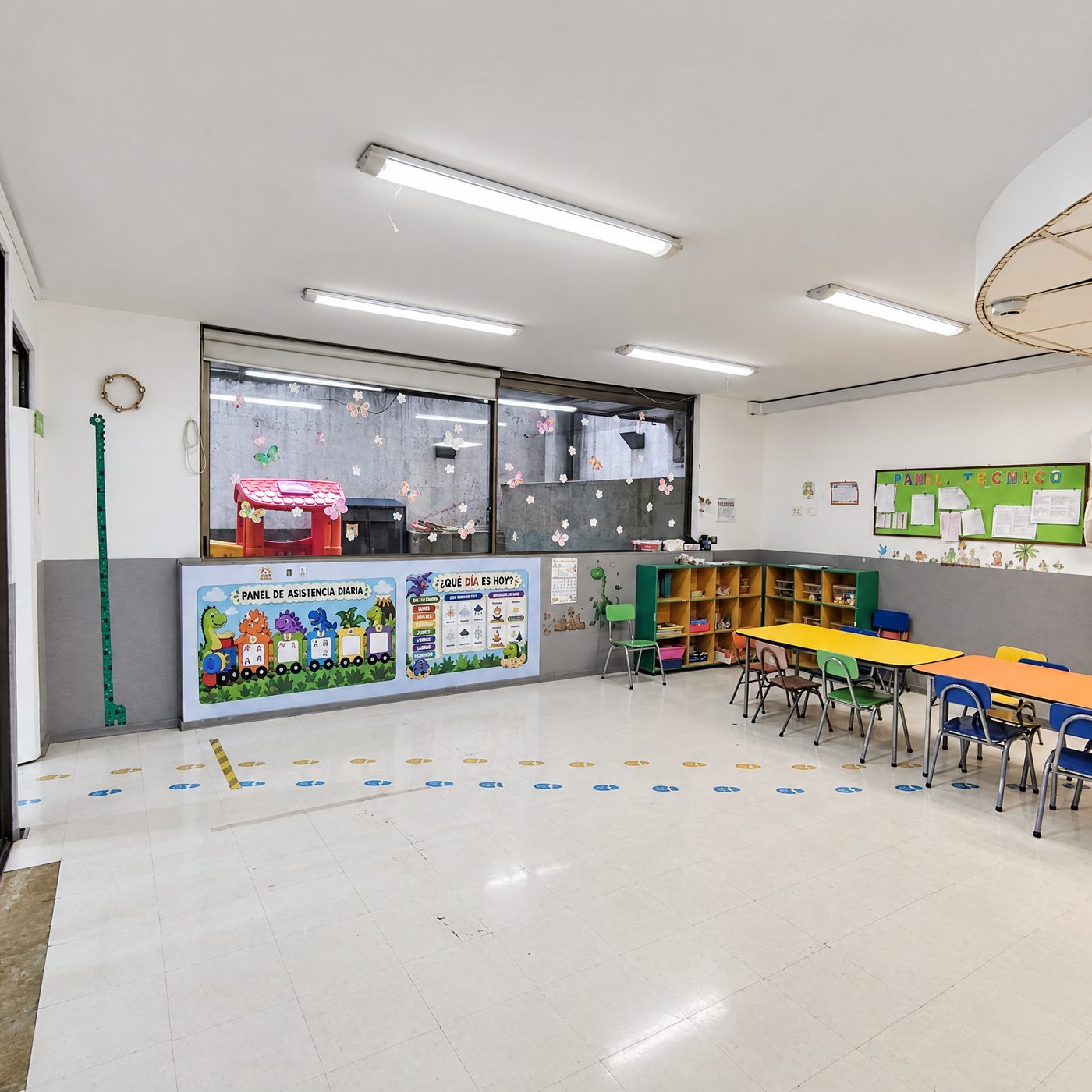 Sala del jardín infantil con mesas de colores, sillas pequeñas y panel de asistencia